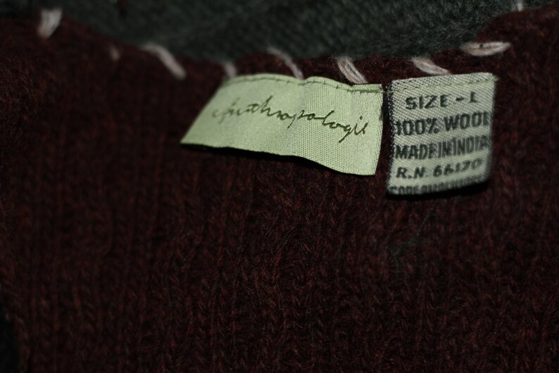 K&ouml;nnte beinhalten: Ein gr&uuml;nes Etikett mit dem Text "anthropologie" und ein braunes Etikett mit dem Text "SIZE - L 100% WOOL MADE IN INDIA R.N. 66170" befestigt an einem dunkelbraunen Strickstoff.