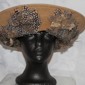 Op de afbeelding: Een brede, bruine vilt hoed met een decoratieve band van bruine en witte veren.