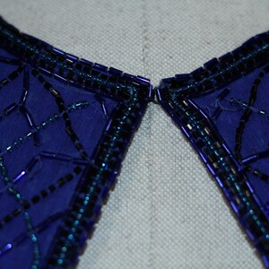 May include: Close-up of a blue fabric collar with a beaded trim. The beads are arranged in a geometric pattern and are a mix of black and iridescent blue.