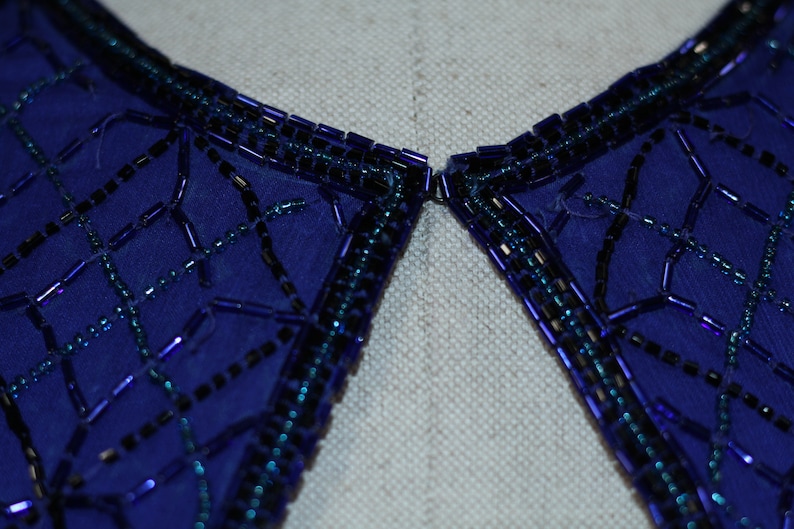 May include: Close-up of a blue fabric collar with a beaded trim. The beads are arranged in a geometric pattern and are a mix of black and iridescent blue.