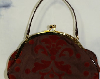 May include: A brown handbag with a red velvet floral pattern and a gold clasp. The bag has a brown handle and a shiny, dark brown trim. The bag is hanging from a silver hook.