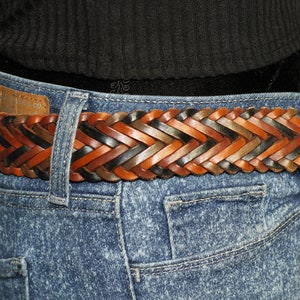 K&ouml;nnte beinhalten: Ein geflochtener Lederg&uuml;rtel mit gewebten Str&auml;ngen in Braun, Schwarz und Tan. Der G&uuml;rtel wird &uuml;ber einer blauen Jeans getragen.