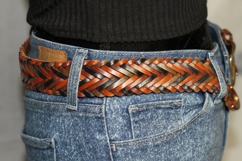 K&ouml;nnte beinhalten: Ein geflochtener Lederg&uuml;rtel mit gewebten Str&auml;ngen in Braun, Schwarz und Tan. Der G&uuml;rtel wird &uuml;ber einer blauen Jeans getragen.