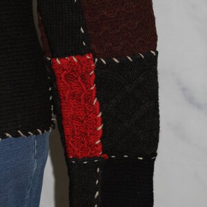 K&ouml;nnte beinhalten: Nahaufnahme eines rot, schwarz und braunen Strickpullovers mit Patchwork-Muster. Der Pullover hat ein Zopfmuster und ist mit wei&szlig;em Garn zusammengen&auml;ht.