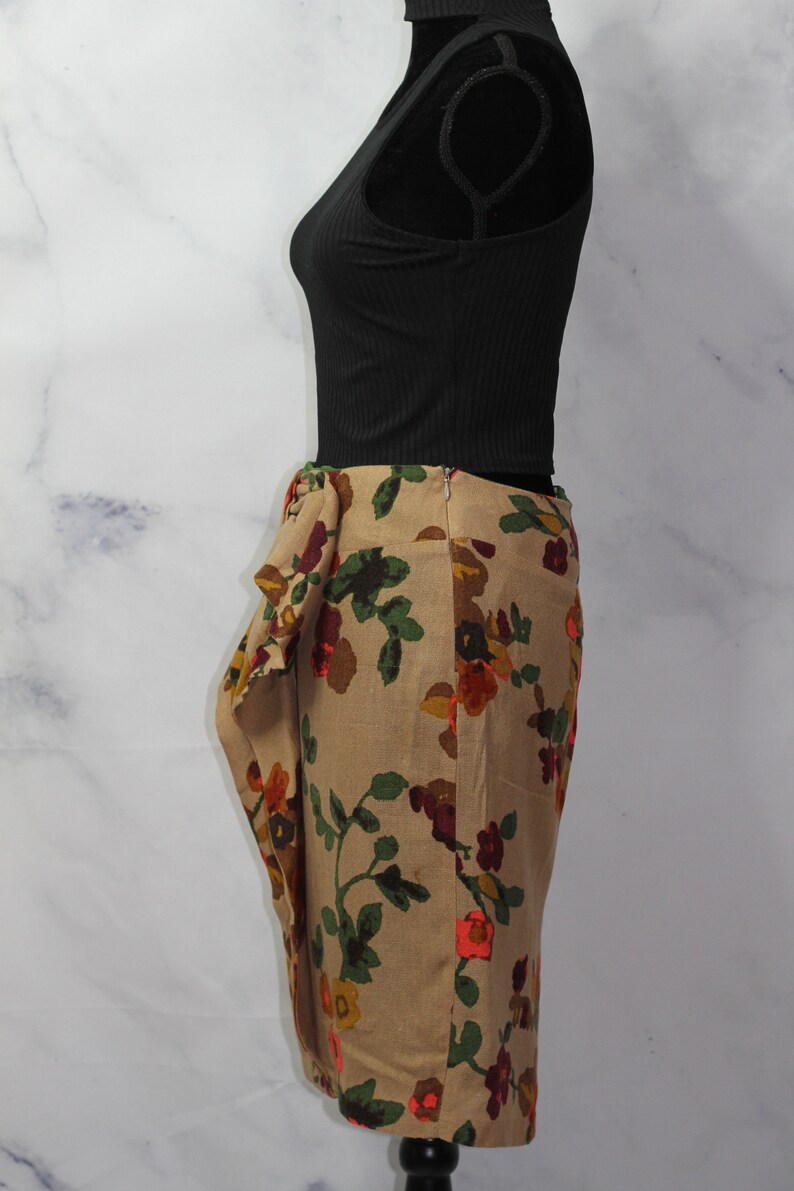 May include: A beige skirt with a floral print featuring red, orange, and green flowers. The skirt has a gathered detail at the waist and a fitted silhouette.