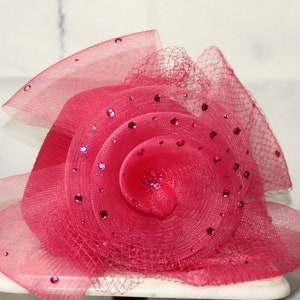 May include: A pink fabric flower hat with a swirl design and rhinestones.