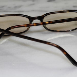 Puede incluir: Gafas de pasta de carey con monturas marrones y lentes transparentes. Las gafas están sobre una superficie de mármol blanco.