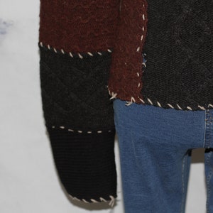 K&ouml;nnte beinhalten: Ein brauner, schwarzer und cremefarbener Patchwork-Pullover mit einem Zopfmuster. Der Pullover ist mit hochgekrempelten &Auml;rmeln und einem ausgefransten Saum zu sehen.