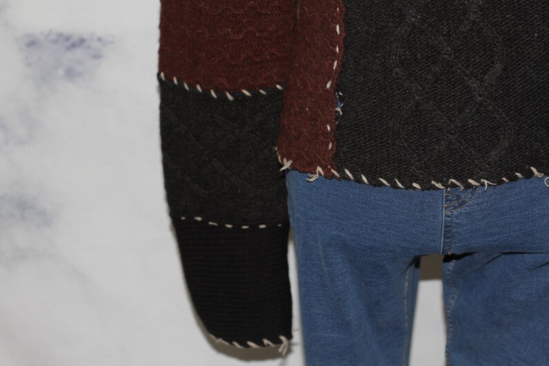 K&ouml;nnte beinhalten: Ein brauner, schwarzer und cremefarbener Patchwork-Pullover mit einem Zopfmuster. Der Pullover ist mit hochgekrempelten &Auml;rmeln und einem ausgefransten Saum zu sehen.