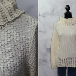 Op de afbeelding: Crèmekleurige trui met een textuurgebreid patroon. De trui heeft een hoge, gerimpelde kraag en lange mouwen. De onderste zoom heeft een decoratief opengewerkt ontwerp. De trui wordt op een mannequin gepresenteerd tegen een lichte achtergrond.