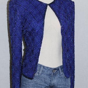 May include: A royal blue sequined jacket with a black trim and open front. The jacket is made of a soft, silky fabric and has a delicate, intricate design.