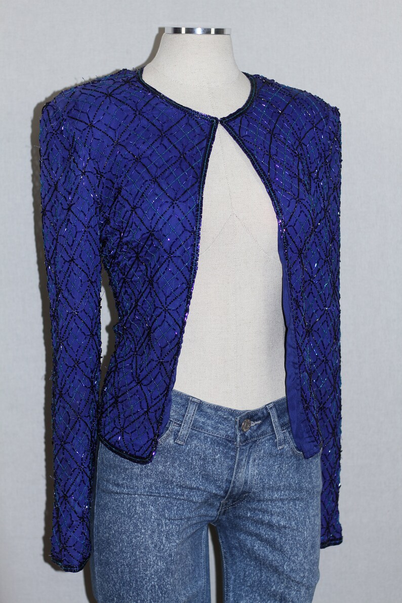 May include: A royal blue sequined jacket with a black trim and open front. The jacket is made of a soft, silky fabric and has a delicate, intricate design.