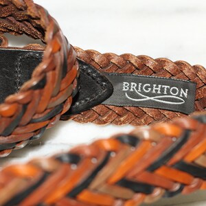 K&ouml;nnte beinhalten: Ein geflochtener Lederg&uuml;rtel mit einer schwarzen Lederschnalle und einem braunen Lederanh&auml;nger mit der Aufschrift "BRIGHTON" in wei&szlig;en Buchstaben.