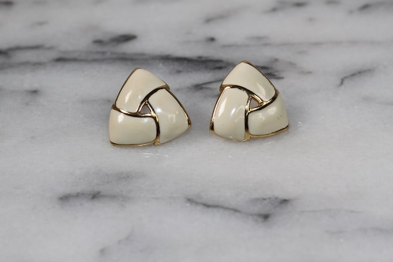 May include: A pair of gold tone earrings with a white enamel design. The earrings are shaped like a triangle and have a woven pattern.
