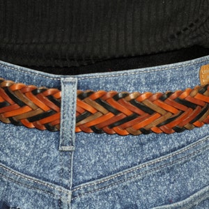 K&ouml;nnte beinhalten: Ein geflochtener Lederg&uuml;rtel mit gewebten Str&auml;ngen in Braun, Schwarz und Tan. Der G&uuml;rtel wird &uuml;ber einer blauen Jeans getragen.