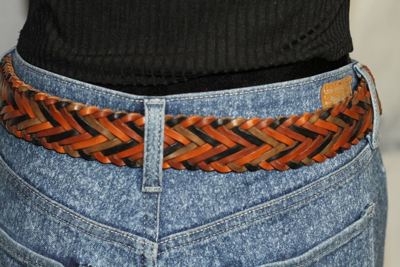 K&ouml;nnte beinhalten: Ein geflochtener Lederg&uuml;rtel mit gewebten Str&auml;ngen in Braun, Schwarz und Tan. Der G&uuml;rtel wird &uuml;ber einer blauen Jeans getragen.