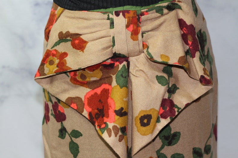 May include: A beige skirt with a large bow in the back. The skirt has a floral print with red, orange, yellow, and green flowers.