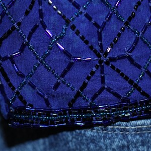 May include: A close-up of a blue denim fabric with a decorative beaded design. The beads are arranged in a geometric pattern, creating a grid-like effect. The beads are a mix of dark blue, light blue, and purple.
