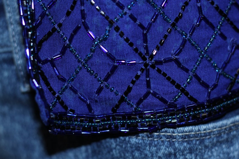 May include: A close-up of a blue denim fabric with a decorative beaded design. The beads are arranged in a geometric pattern, creating a grid-like effect. The beads are a mix of dark blue, light blue, and purple.
