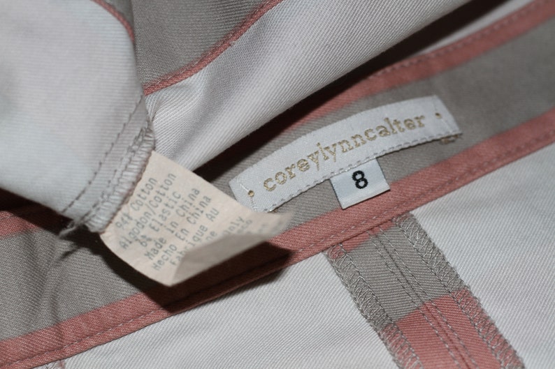 Puede incluir: Primer plano de una etiqueta de prenda con el texto "coreyiynncalter" y el n&uacute;mero "8" impreso en ella. La etiqueta est&aacute; unida a una tela de rayas grises claras y rosas.
