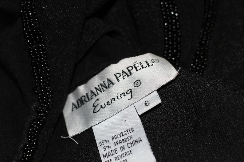 Pode incluir: Uma etiqueta branca com texto preto que diz "ADRIANNA PAPELL Evening" e "95% POLYESTER 5% SPANDEX MADE IN CHINA OFF REVERSE DE" com uma etiqueta de tamanho 6 anexada.
