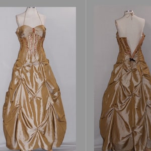 Puede incluir: Un vestido dorado sin tirantes con un corpiño de cuentas y una falda con volantes. El vestido tiene la espalda baja y está ajustado en la cintura con una cinta negra.