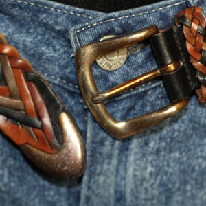 K&ouml;nnte beinhalten: Nahaufnahme eines geflochtenen Lederg&uuml;rtels mit einer Messingschnalle. Der G&uuml;rtel ist um eine blaue Jeans geschnallt.