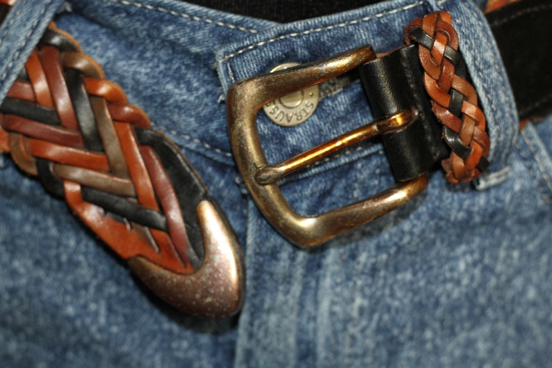 K&ouml;nnte beinhalten: Nahaufnahme eines geflochtenen Lederg&uuml;rtels mit einer Messingschnalle. Der G&uuml;rtel ist um eine blaue Jeans geschnallt.