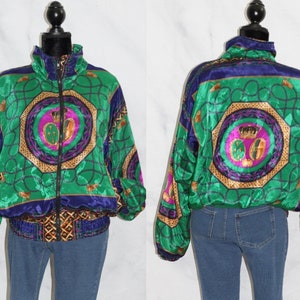 Puede incluir: Una chaqueta de satén verde, azul, dorado y morado con un patrón geométrico. La chaqueta tiene una cremallera y un cuello alto.