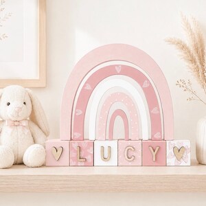 May include: A wooden rainbow stacker in shades of pink and white with heart and dot patterns, positioned behind six wooden blocks spelling the name LUCY with heart accents on the end blocks, accompanied by a plush cream-coloured bunny toy.