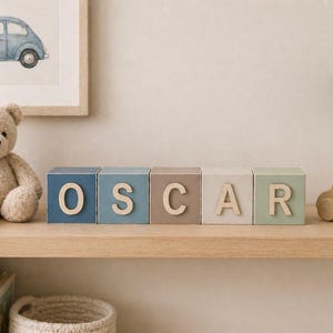 May include: Five square wooden blocks arranged in a row, each painted in a different muted colour including blue, grey, and green. Each block features a large, light-coloured wooden letter attached to the front, collectively spelling the name OSCAR.