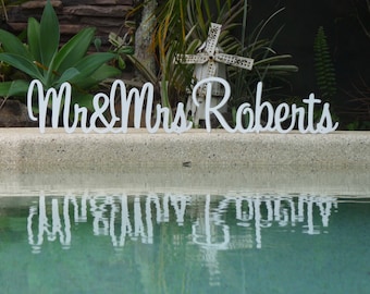 May include: White wooden sign with the text "Mr & Mrs Roberts" in cursive lettering. The sign is placed on a concrete ledge in front of a swimming pool.