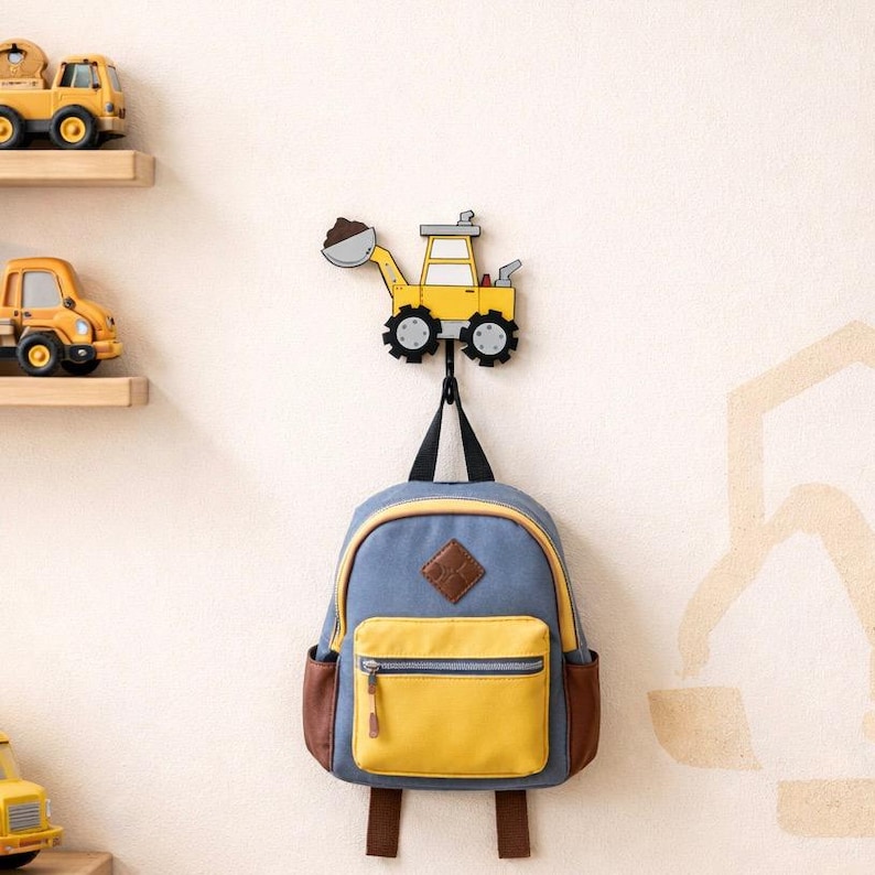 May include: A yellow and black excavator-shaped wall hook with a black hook holding a blue and yellow backpack. The backpack has brown accents and a front pocket. Toy trucks are on shelves in the background.