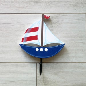 Peut inclure: Un crochet mural décoratif en forme de voilier. Le bateau a une coque bleue, des voiles blanches avec des rayures rouges et un mât marron avec un drapeau rouge. Trois cercles blancs sont sur la coque. Un crochet noir s'étend du bas.