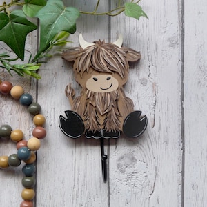 Peut inclure: Un crochet mural en bois en forme de vache des Highlands, dans des tons de marron, beige et noir. La vache a un visage souriant, des cornes blanches et une longue robe hirsute. Le crochet est noir et s'étend du ventre de la vache. Des perles en bois décoratives et de la verdure sont en arrière-plan.