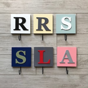 May include: Six square wooden wall hooks, each with a different letter in a bold font. The letters are black, gray, white, red, and green. The background colors are white, yellow, blue, gray, and pink. Each hook has a black metal hook.