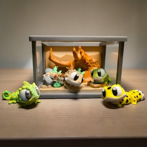 May include: A collection of four cartoon gecko figurines in various colors, including green, yellow, and brown, with large, expressive eyes. One is outside the glass terrarium, while the others are inside, near a decorative branch.