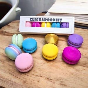 Könnte beinhalten: Ein Set bunter Macarons in einer weißen Box mit dem Text "Clickaroonies". Die Macarons sind in einer Reihe angeordnet und in den Farben Rosa, Blau, Gelb, Grün und Lila.