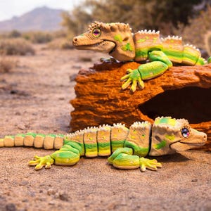 Peut inclure: Deux jouets articulés en forme de lézard, dans des tons de vert, jaune et beige, reposent sur une bûche et du sable bruns et texturés. Les lézards ont des détails précis, notamment des corps segmentés et des yeux expressifs. L'arrière-plan montre un paysage désertique avec des cactus.