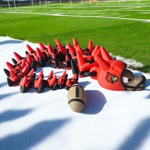 May include: A red and black dragon-shaped training aid with a football. The dragon has a helmet with a logo. The football is tan with black stitching. The items are on a white surface with a green field in the background.
