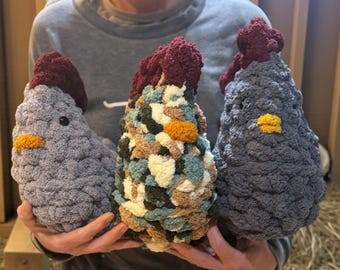 May include: Three handmade crochet chicken plushies with textured yarn. Two chickens are solid gray, and one is a variegated mix of cream, tan, teal, and dark green. Each features a dark red textured comb, a small yellow beak, and black eyes.