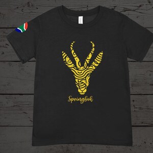 Puede incluir: Camiseta negra con un dise&ntilde;o de Springbok amarillo. El gr&aacute;fico representa una cabeza de ant&iacute;lope estilizada con patrones detallados y la palabra "Springbok" en una fuente cursiva. Una peque&ntilde;a bandera sudafricana est&aacute; en la manga izquierda.