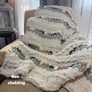 May include: A cream-colored, chunky knit blanket with stripes of blue, brown, and black. The blanket is draped over a chair. The words "Non-shedding" are written on the blanket.