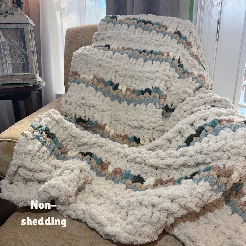 May include: A cream-colored, chunky knit blanket with stripes of blue, brown, and black. The blanket is draped over a chair. The words "Non-shedding" are written on the blanket.