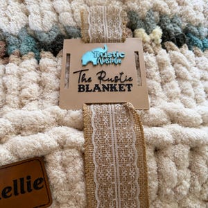 May include: A cream-colored, chunky knit blanket with a tag that reads "The Rustic Blanket." The blanket is tied with a burlap ribbon with a white lace trim. A small brown leather tag is also attached.