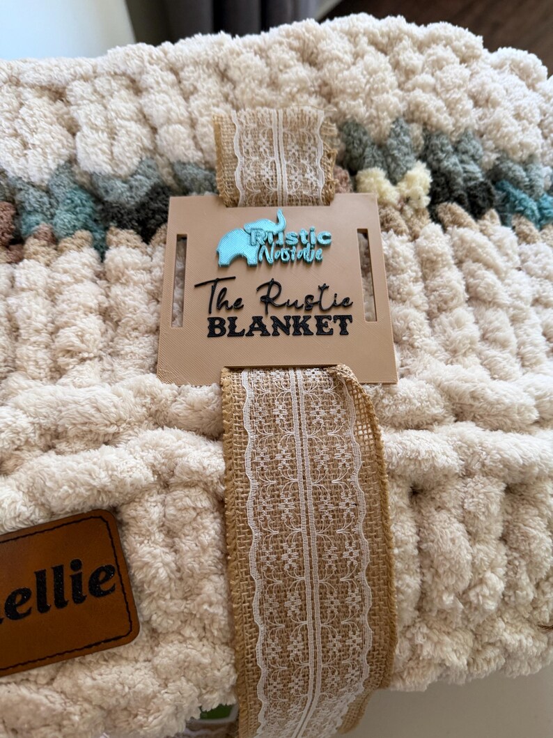 May include: A cream-colored, chunky knit blanket with a tag that reads "The Rustic Blanket." The blanket is tied with a burlap ribbon with a white lace trim. A small brown leather tag is also attached.