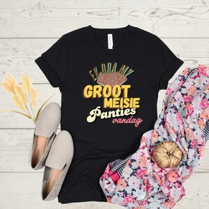 May include: Black V-neck t-shirt with the text "Ek dra my groot meisie panties vandag" in yellow, brown, and white. The shirt is paired with gray slip-on shoes and a floral scarf.