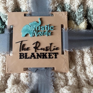 May include: A plush, cream-colored blanket with a textured pattern, tied with a blue ribbon. A tan tag with the text "Rustic Nostalgie" and "The Rustic BLANKET" is attached to the blanket.