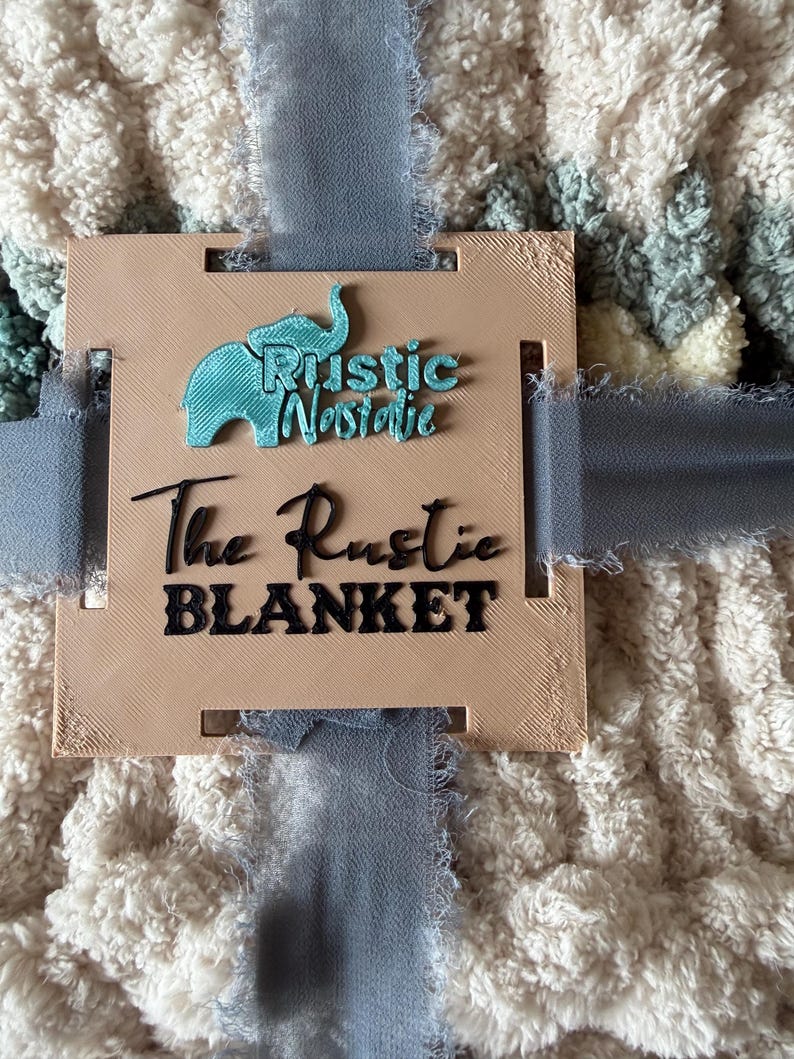 May include: A plush, cream-colored blanket with a textured pattern, tied with a blue ribbon. A tan tag with the text "Rustic Nostalgie" and "The Rustic BLANKET" is attached to the blanket.