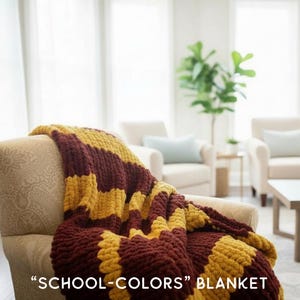 May include: A chunky knit blanket in maroon and gold stripes is draped over a beige armchair. The blanket features the text "SCHOOL-COLORS" BLANKET. The cozy throw is a decorative accent for a living room.